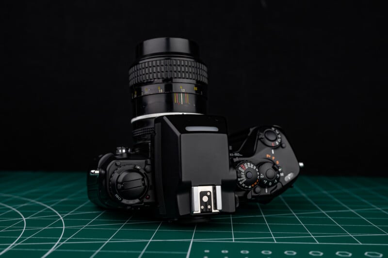 A black vintage film camera with a lens, viewed from above, sits on a green cutting mat with white grid lines, against a black background.