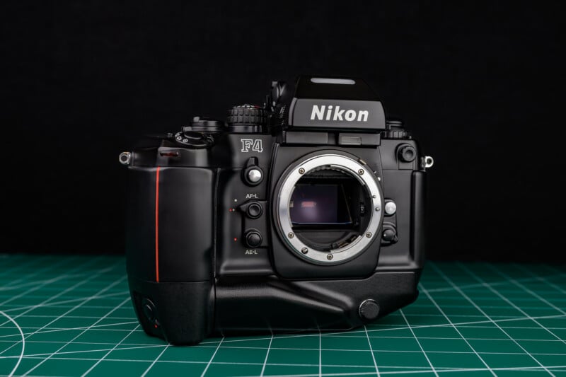 A Nikon F4 film camera body without a lens is placed on a green cutting mat with a black background. The camera's lens mount and internal mirror are visible.