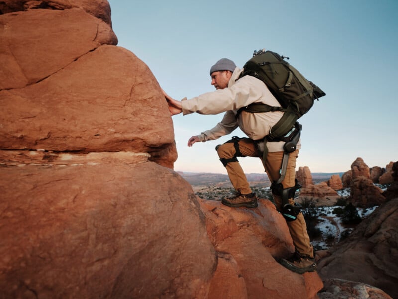 A person wearing a backpack and a gray beanie climbs a large red rock formation in a desert landscape with scattered rock towers and a clear sky in the background.