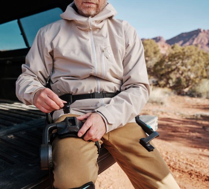 A person wearing a light jacket and brown pants sits on a truck bed outdoors, adjusting a black knee brace on their leg in a rocky, desert-like landscape.