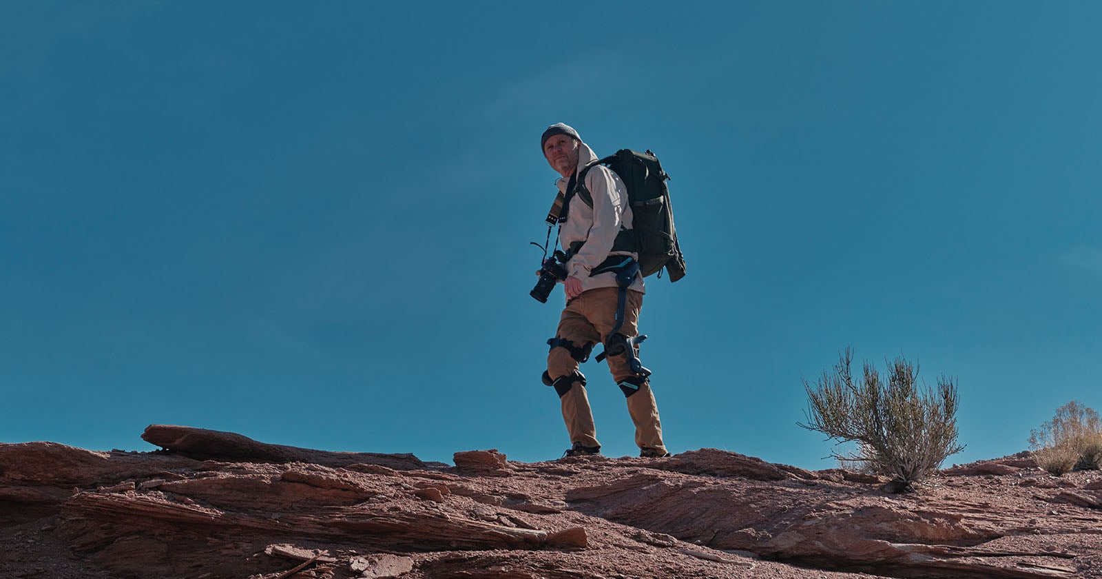 A person wearing outdoor gear and a backpack stands on a rocky terrain under a clear blue sky, holding a camera and looking off into the distance. Sparse desert vegetation is visible nearby.