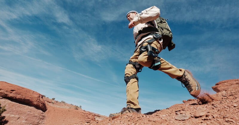 A person wearing outdoor gear and a backpack hikes up a rocky hill under a clear blue sky, with a close-up focus on their legs in motion and dirt kicking up from their boots.