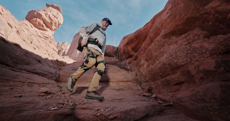 A person wearing hiking gear and a backpack walks through a rocky desert canyon, surrounded by red sandstone cliffs under a blue sky.