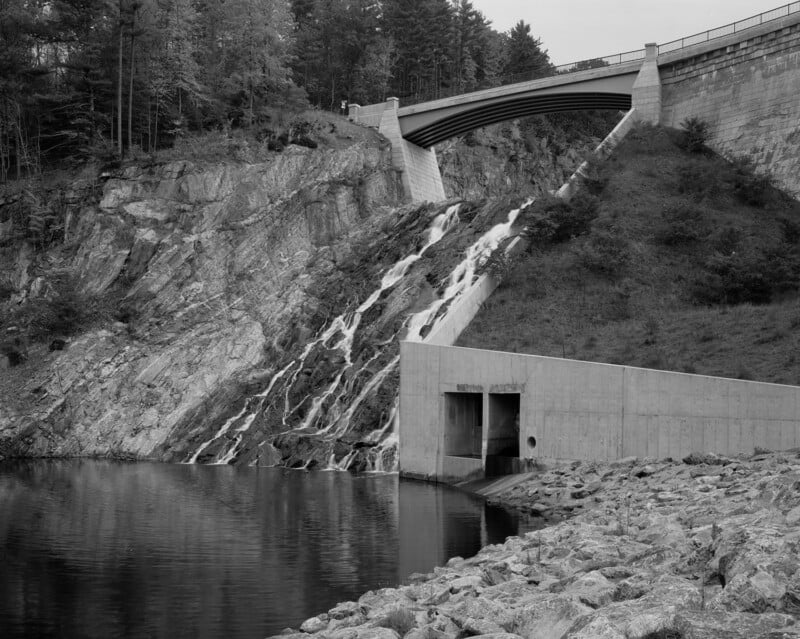 Un puente de presa de hormigón se extiende sobre una ladera rocosa, con agua cayendo en cascada por un aliviadero hacia un tranquilo embalse debajo. La cima de la colina está densamente boscosa y la costa en primer plano está cubierta de rocas. La imagen está en blanco y negro.