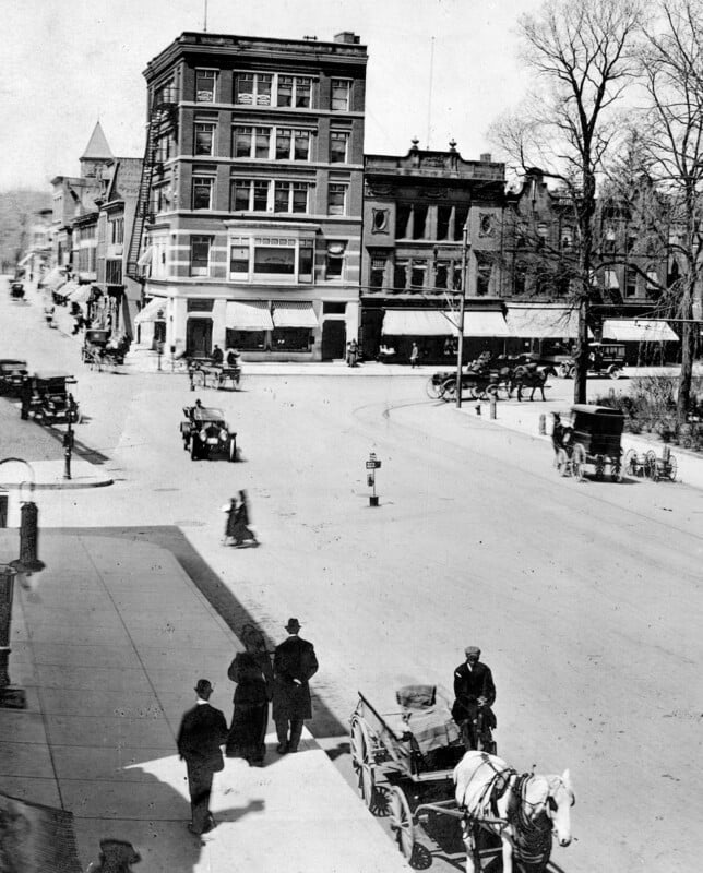 Esta histórica fotografía en blanco y negro muestra una calle de la ciudad con carruajes tirados por caballos, los primeros automóviles y gente caminando por la acera cerca de edificios de ladrillo de varios pisos y escaparates. Las calles están bordeadas de árboles desnudos.