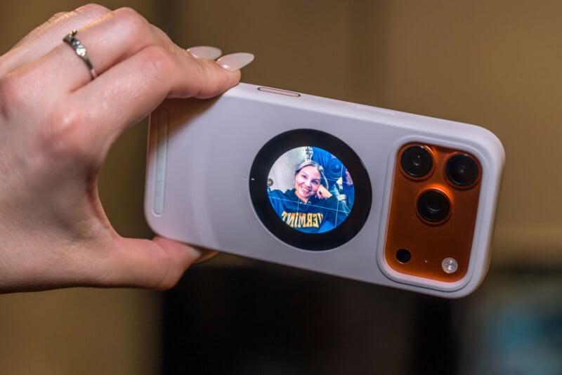 Una mano sostiene un teléfono inteligente con una pantalla circular en la parte posterior que muestra a un hombre sonriente tomándose una selfie. El teléfono tiene una carcasa blanca y tres lentes de cámara visibles.