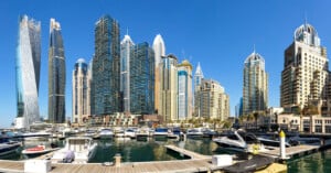 A panoramic view of a marina with many yachts docked in front of modern, tall skyscrapers under a clear blue sky, showcasing an urban waterfront cityscape.