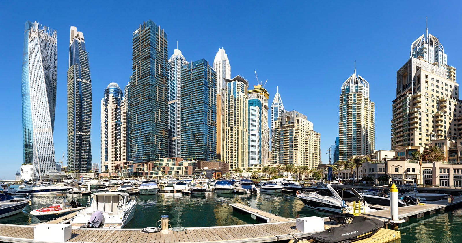 A panoramic view of a marina with many yachts docked in front of modern, tall skyscrapers under a clear blue sky, showcasing an urban waterfront cityscape.