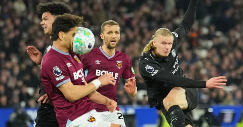A soccer player in black kicks the ball as two West Ham players in maroon attempt to block, with one taking the ball to the face. The stadium is full of spectators in the background.