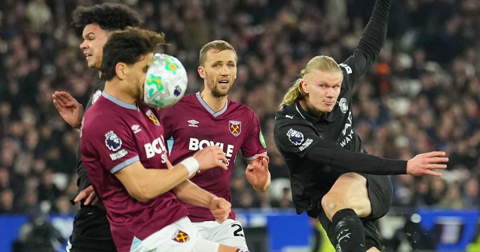 A soccer player in black kicks the ball as two West Ham players in maroon attempt to block, with one taking the ball to the face. The stadium is full of spectators in the background.
