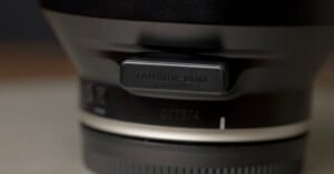 Close-up shot of a camera lens, focusing on a rectangular port cover labeled "TAMRON-LINK." The background is blurred, highlighting the lens details and branding.