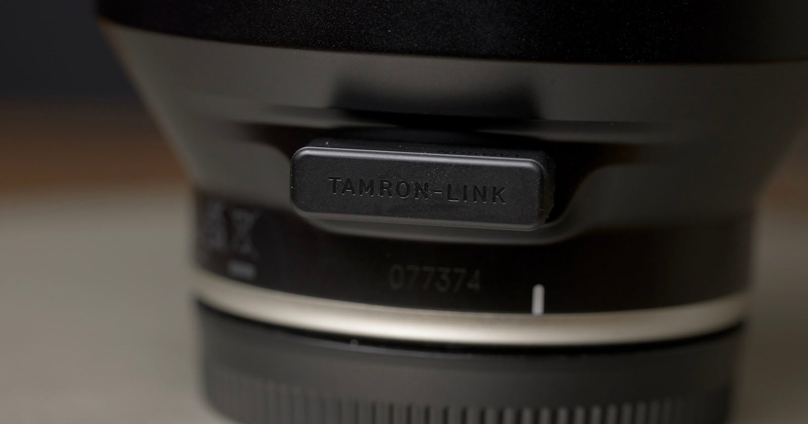 Close-up shot of a camera lens, focusing on a rectangular port cover labeled "TAMRON-LINK." The background is blurred, highlighting the lens details and branding.