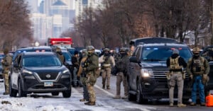 Police officers in tactical gear stand near several police vehicles with flashing lights on a snow-lined street in a city, with other emergency vehicles and high-rise buildings visible in the background.