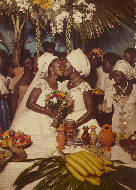 Two women in white dresses and headwraps share a kiss at a wedding, surrounded by guests in colorful attire. A table with fruit and pottery is in the foreground, and a floral arch is in the background.