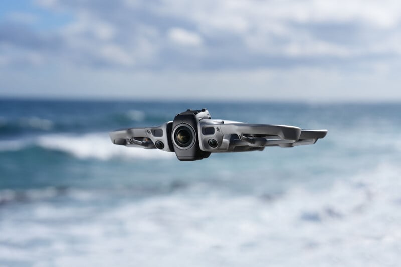 A gray drone flies in midair over the ocean, with waves and a cloudy sky in the background. The drone’s camera is facing forward, capturing the seascape.