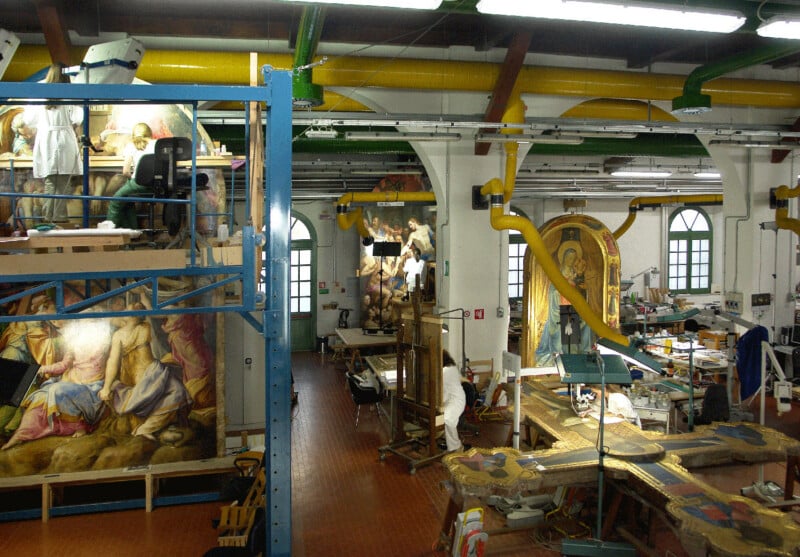 Gran estudio de restauración de arte con techos altos repleto de equipos de conservación, grandes pinturas religiosas y artefactos en restauración. Dos personas trabajan en andamios y mesas entre obras de arte y herramientas de restauración.