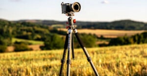 A camera mounted on a tripod stands in a sunlit field with rolling hills and trees blurred in the background, under a clear sky.