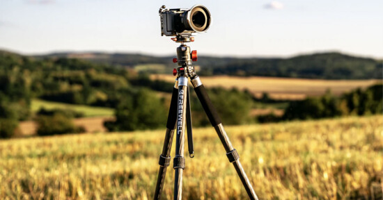 A camera mounted on a tripod stands in a sunlit field with rolling hills and trees blurred in the background, under a clear sky.