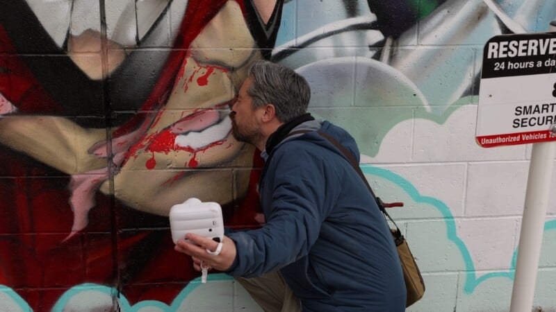 Un hombre con una chaqueta azul, sosteniendo una cámara o dispositivo blanco en una mano y una cámara o dispositivo blanco en la otra, finge en broma besar los labios de una cara pintada en una pared cubierta de graffiti. uno "reservado" A la derecha se ve una señal de stop.