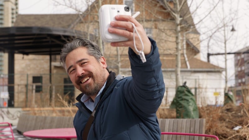 Un hombre sonriente, barbudo y de cabello oscuro se toma una selfie con una cámara instantánea blanca al aire libre, parado frente a un banco y árboles con edificios al fondo.