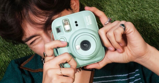 A person with dark hair holds a mint green Instax Mini 9 instant camera up to their eye, appearing to take a photo, while lying on green grass. The person wears rings and a partially open shirt.