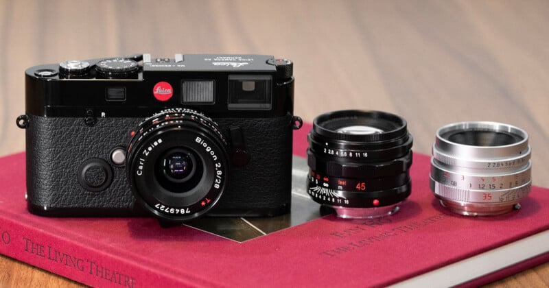 A black Leica camera with a Zeiss lens is placed on a red book, next to two additional camera lenses, one black and one silver, on a wooden surface.