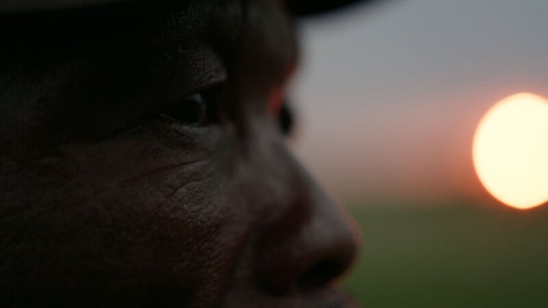 Close-up of a person's face in profile, focusing on their eye and textured skin. A bright, out-of-focus light, possibly the setting sun, glows in the blurred background.