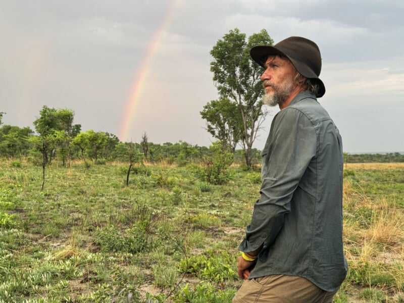 A man in a wide-brimmed hat and green shirt stands in a grassy field, looking into the distance. There is a faint rainbow in the cloudy sky behind him and trees scattered across the landscape.
