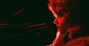 A person in profile faces right, wearing a furry hat and coat, illuminated by red light. Bright red streaks or sparks move across the dark background on the left side of the image.