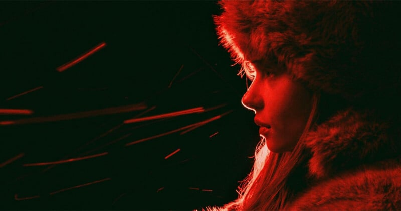 A person in profile faces right, wearing a furry hat and coat, illuminated by red light. Bright red streaks or sparks move across the dark background on the left side of the image.