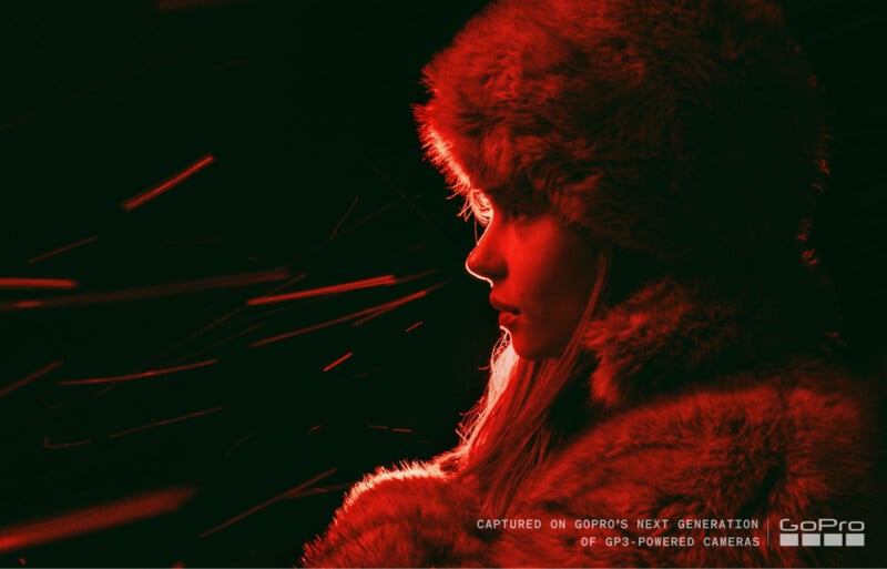 Una mujer que lleva un sombrero y un abrigo peludo está iluminada por una espectacular iluminación roja, con rayos de luz que la rodean. El texto en la parte inferior dice 