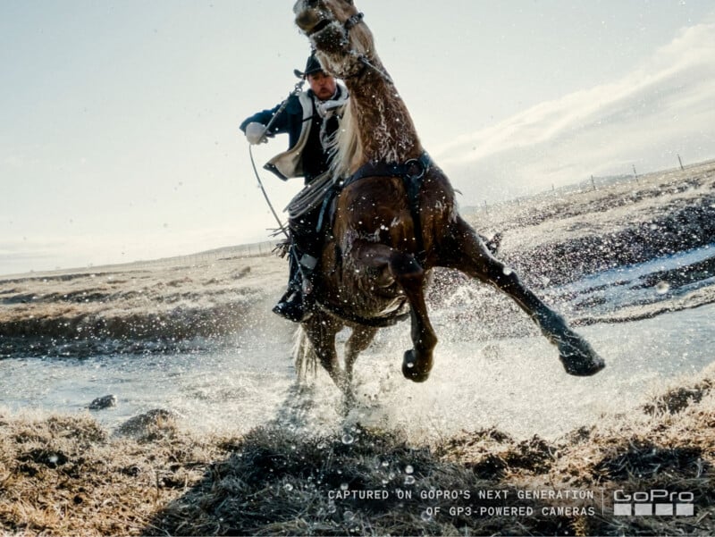 Un hombre vestido con equipo para actividades al aire libre monta un caballo marrón que galopa a través de un arroyo poco profundo, salpicando agua, con un paisaje abierto al fondo. El texto en la esquina menciona la cámara GP3 de GoPro.