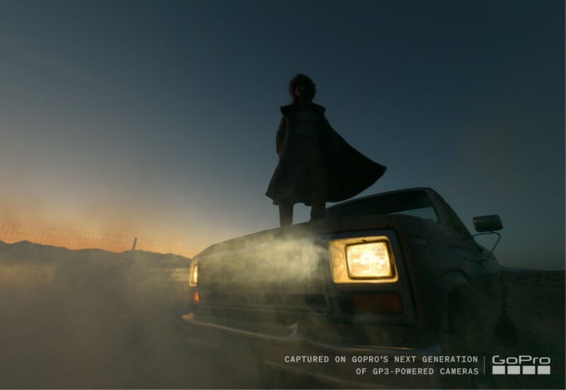 Al anochecer, una persona se encuentra en el techo de un automóvil, reflejando el cielo nocturno. Los faros están encendidos y la niebla o el humo rodean el coche. El texto de la imagen dice: "Tomada con la cámara GP3 de nueva generación de GoPro.