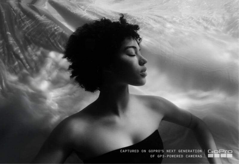 Una mujer de pelo rizado flota bajo el agua con los ojos cerrados, bajo una iluminación tenue. Esta imagen es en blanco y negro y contiene la marca GoPro y texto sobre la cámara GP3 de próxima generación.