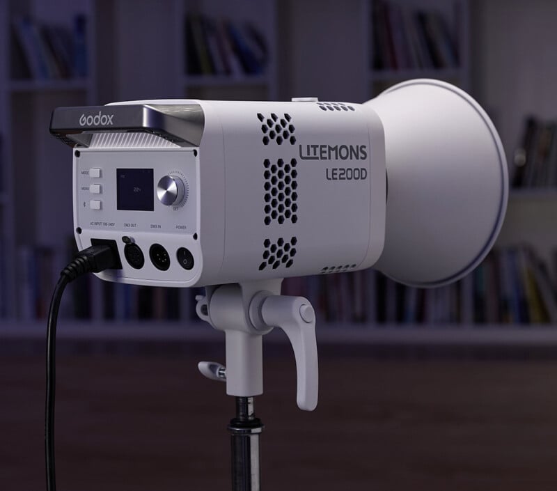 Primer plano de la lámpara de estudio Godox LITEMONS LE200D colocada sobre un soporte, en el interior, con estanterías y libros borrosos en el fondo. El panel de control de la luz y los puertos de conexión son visibles.
