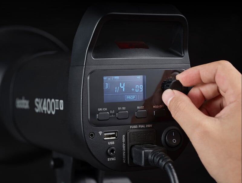 Ajuste la perilla de control del flash de estudio Godox SK400II con una mano y la pantalla digital mostrará la configuración del flash, así como los distintos botones, puertos y etiquetas visibles en el dispositivo.