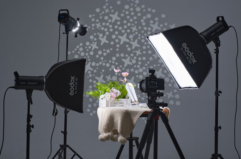 Estudio de fotografía con tres luces softbox, cámara sobre trípode y una pequeña mesa con flores, un libro y adornos sobre un fondo gris con un patrón decorativo blanco.