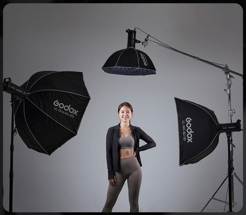 Una mujer en ropa deportiva se encuentra de pie y sonríe en un estudio de fotografía, rodeada por tres grandes luces de softbox Godox en las gradas sobre un fondo gris sólido.