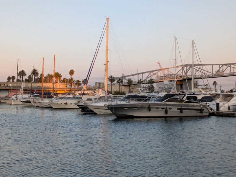 Boats and yachts docked at a marina during sunset, with a pedestrian bridge and palm trees in the background under a clear sky.