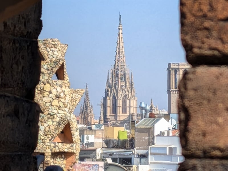 A distant view of a tall Gothic cathedral spire rising above city rooftops, framed by the stone walls and textured surfaces of nearby buildings.