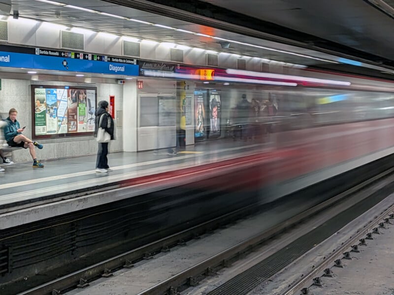 A subway train speeds past the Diagonal station platform in Barcelona, creating a motion blur. A few people wait on the platform, some seated and some standing, with advertisements and signs visible on the walls.