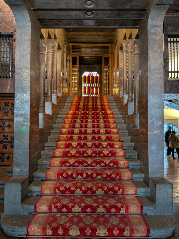 A grand stone staircase with a red and gold patterned carpet runner leads up to stained glass windows in a historic, ornate building. Two people stand off to the side near the entrance.