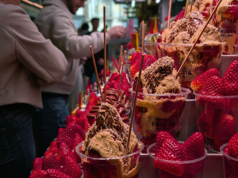 Plastic cups filled with fresh strawberries and chocolate-drizzled desserts are displayed on a market stand, with people in the background selecting items.