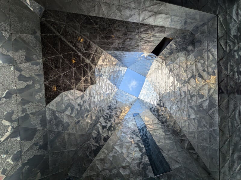 View looking up through a geometric, mirrored architectural structure with reflective silver surfaces and an opening at the top, revealing blue sky and clouds.