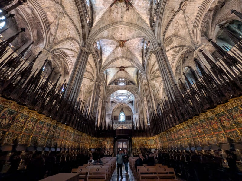 A wide-angle view of a gothic cathedral’s ornate interior, featuring high vaulted ceilings, pointed arches, intricate stonework, and detailed wooden choir stalls lining both sides, with visitors standing in the central aisle.