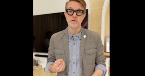 A man with short light hair, wearing black glasses, a gray jacket, and a light blue button-up shirt, gestures with one hand while standing indoors in front of a TV and bookshelves.