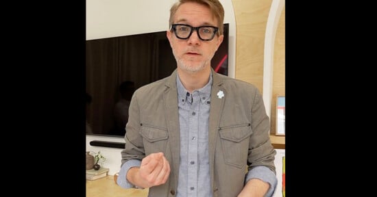 A man with short light hair, wearing black glasses, a gray jacket, and a light blue button-up shirt, gestures with one hand while standing indoors in front of a TV and bookshelves.