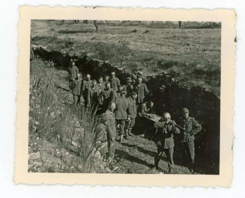 A group of soldiers in uniform stand in and near a trench in a grassy field, with some facing the camera and others looking away, in a black-and-white vintage photograph.