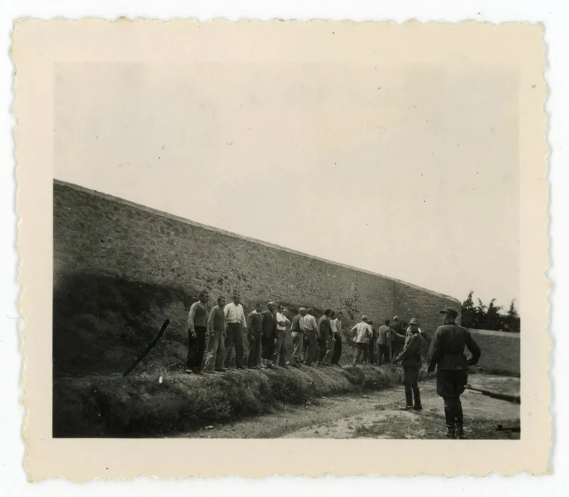 A group of men stands facing a stone wall with their backs turned, while two uniformed soldiers stand nearby, outdoors on a dirt area. The scene appears tense and somber.