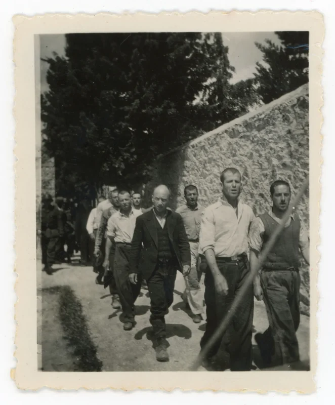 A group of men walk in a line along a narrow, tree-lined path next to a stone wall. The men wear plain clothing and have serious expressions. The photo is black and white with scalloped edges.